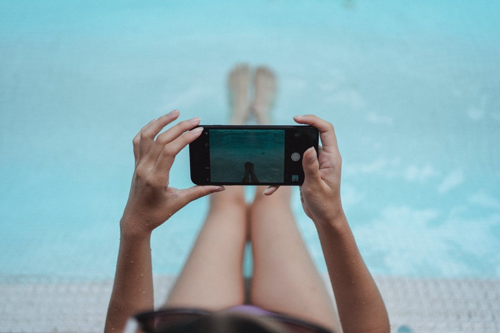 Mulher usando celular perto de uma piscina — Foto: Pexels